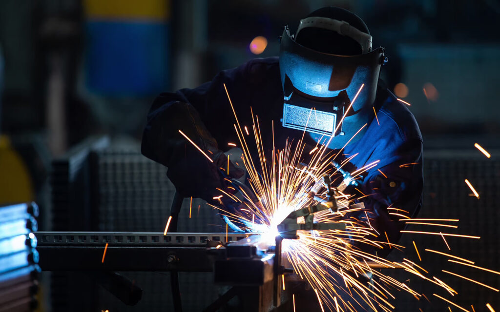 A service tech wearing a welding helmet with sparks flying from welding metal pieces on a table.